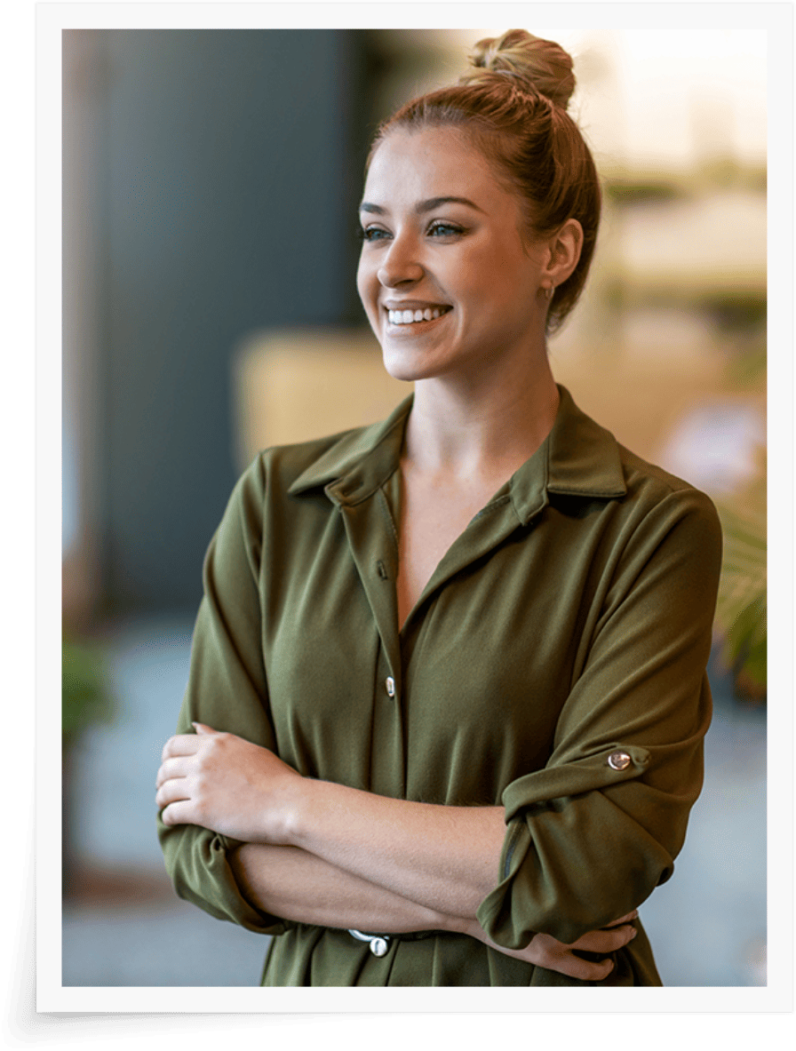 Eine junge, berufstätige Frau mit zu einem Knoten gebundenen Haaren und einer dunkelgrünen Bluse steht lächelnd mit verschränkten Armen da. Sie befindet sich in einem gut beleuchteten Coworking-Raum, wahrscheinlich einem An image from EDGE Workspaces.