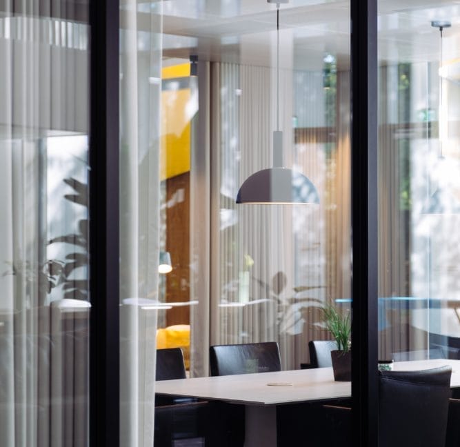 Modern coworking space seen through vertical blinds. The room features a large table with chairs, pendant lights, and plants, framed by floor-to-ceiling windows and reflective surfaces. An image from EDGE Workspaces.