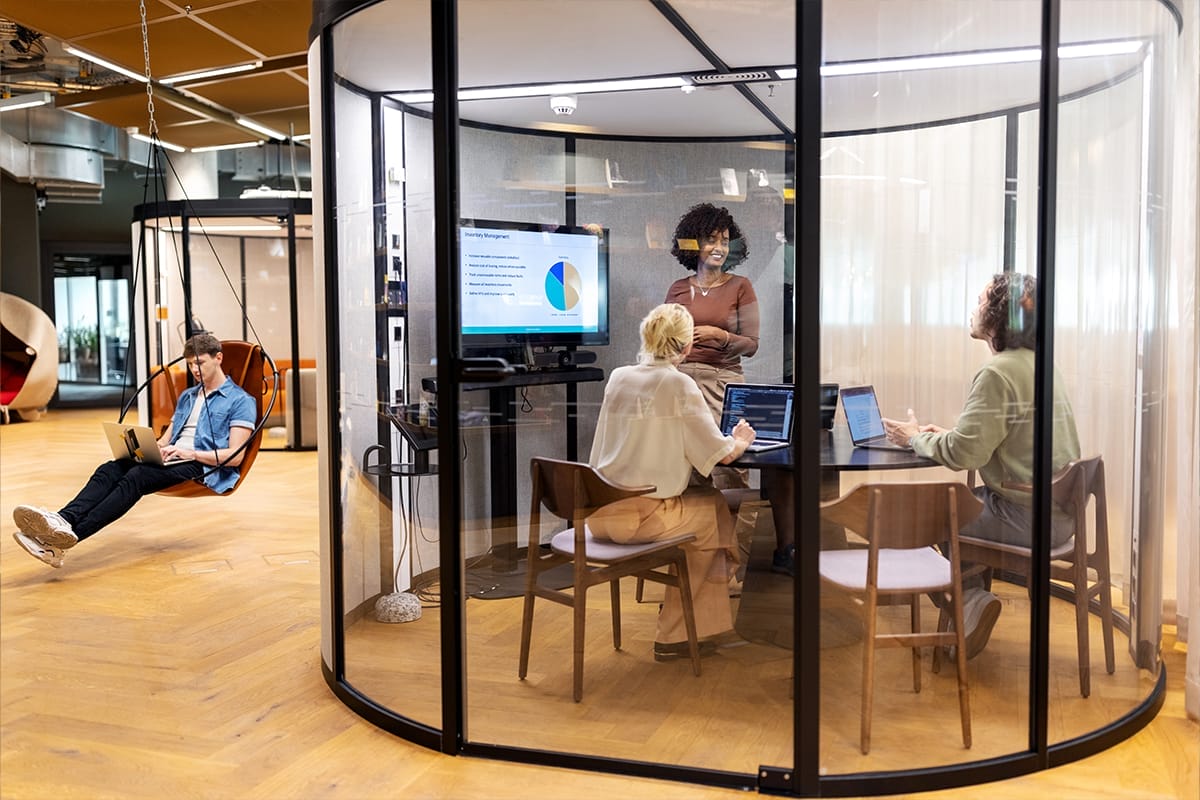 Four diverse professionals in a modern coworking office meeting room with glass walls. A man lounges in a hanging chair with a laptop, while three women engage with a presentation on a screen, one standing An image from EDGE Workspaces.
