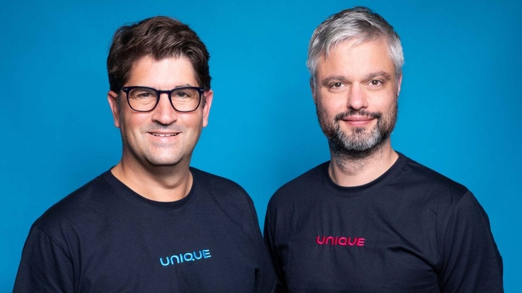 Zwei Männer in schwarzen T-Shirts mit der Aufschrift „unique“ lächeln in die Kamera und stehen vor einem blauen Hintergrund. Der Mann links, der eine Brille trägt und braune Haare hat, scheint ein An image from EDGE Workspaces.