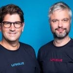 Two smiling males in black t-shirts labeled "unique" standing side by side against a vibrant blue backdrop in a coworking space. The man on the left wears glasses and has dark, wavy hair An image from EDGE Workspaces.