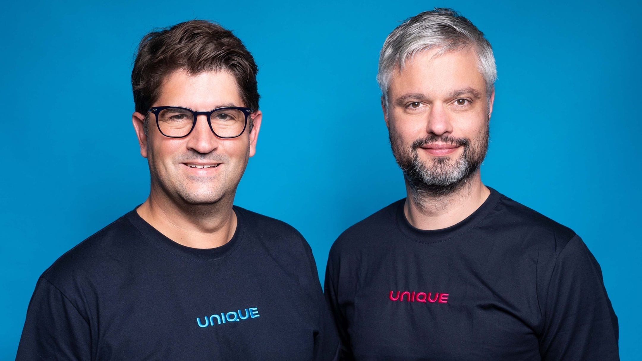 Zwei Männer in schwarzen T-Shirts mit der Aufschrift „unique“ lächeln in die Kamera und stehen vor einem blauen Hintergrund. Der Mann links, der eine Brille trägt und braune Haare hat, scheint ein An image from EDGE Workspaces.